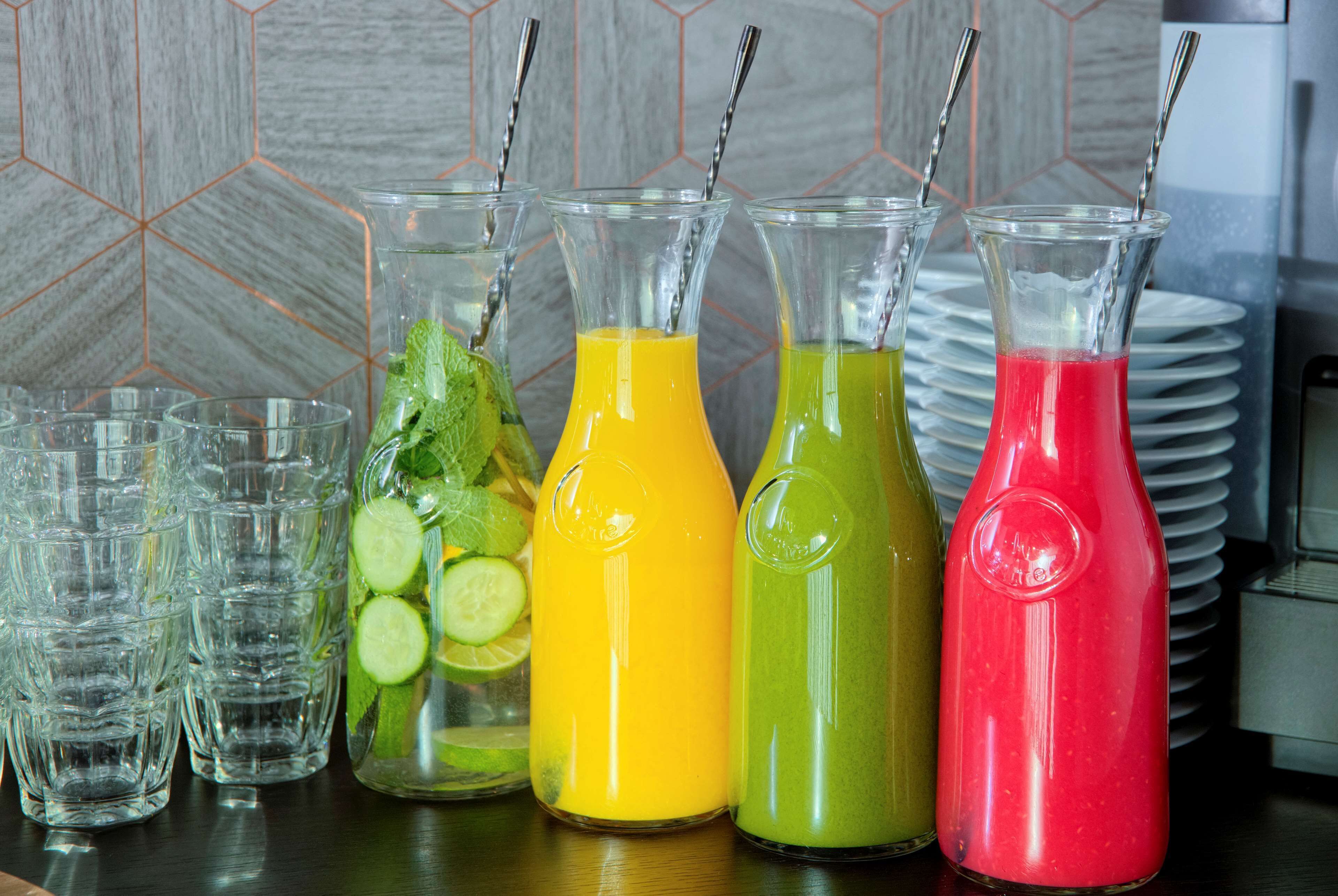 Variedad de bebidas coloridas en botellas de vidrio y vasos, con un backsplash de azulejos al fondo.