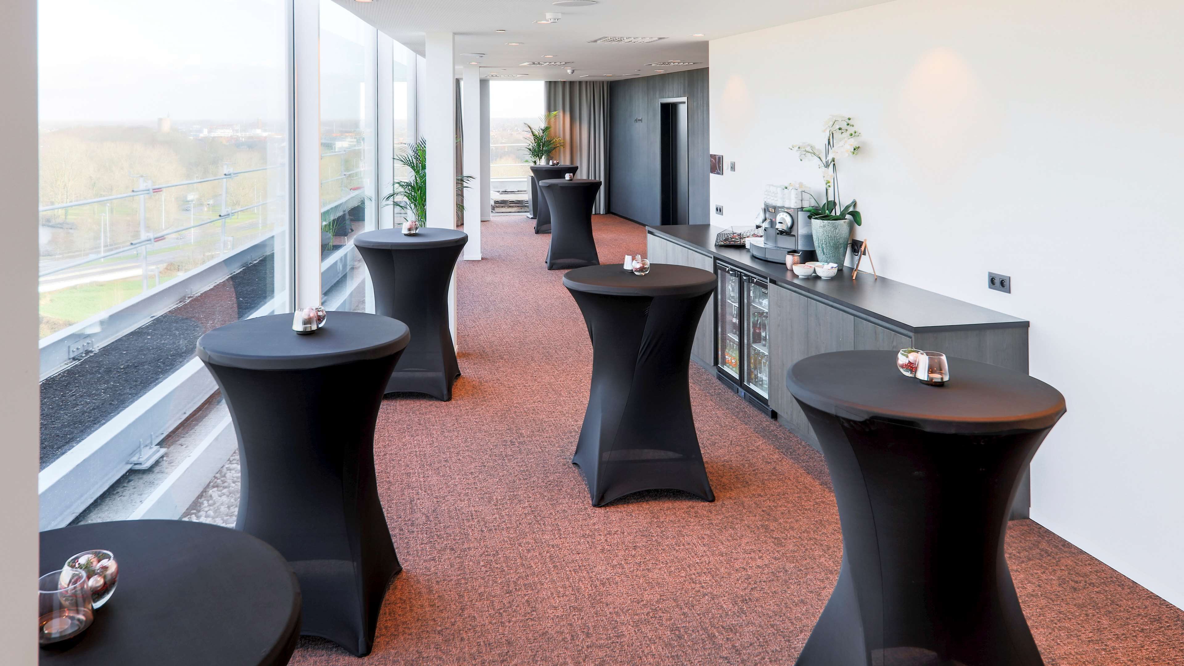 Un vestíbulo de hotel moderno con mesas redondas negras, un mostrador de recepción y amplias ventanas con vistas a la ciudad.