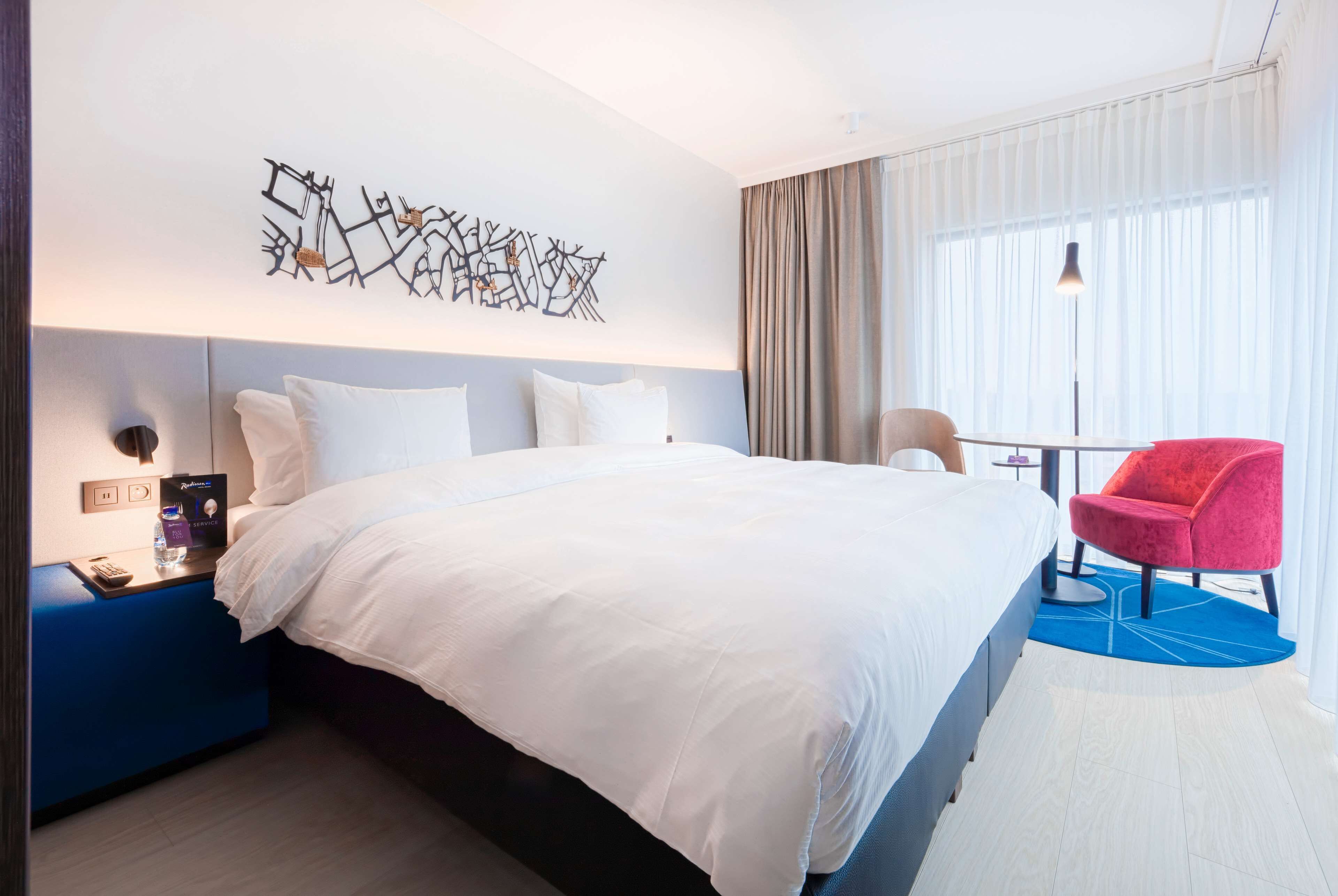 Une chambre d’hôtel moderne avec un grand lit, une table de chevet bleue, une chaise rouge et une œuvre d’art murale.