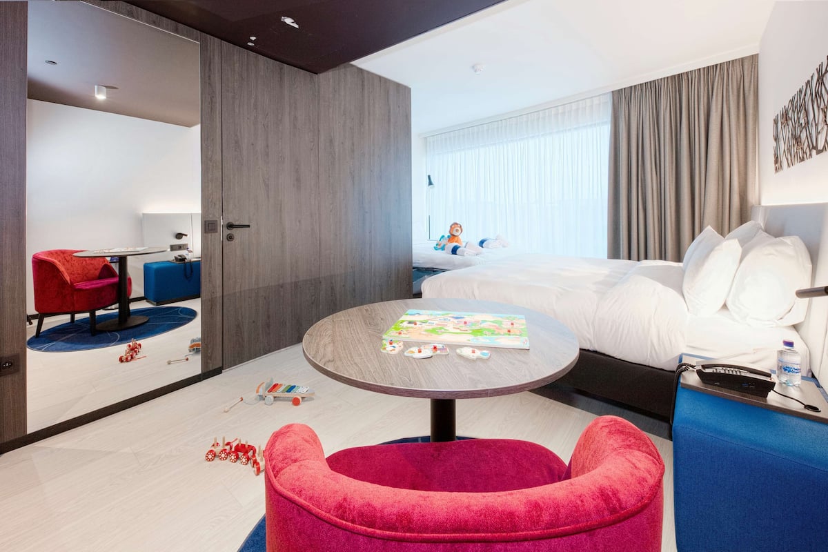 A modern hotel room with a large bed, a round table, a red chair, and a blue sofa.