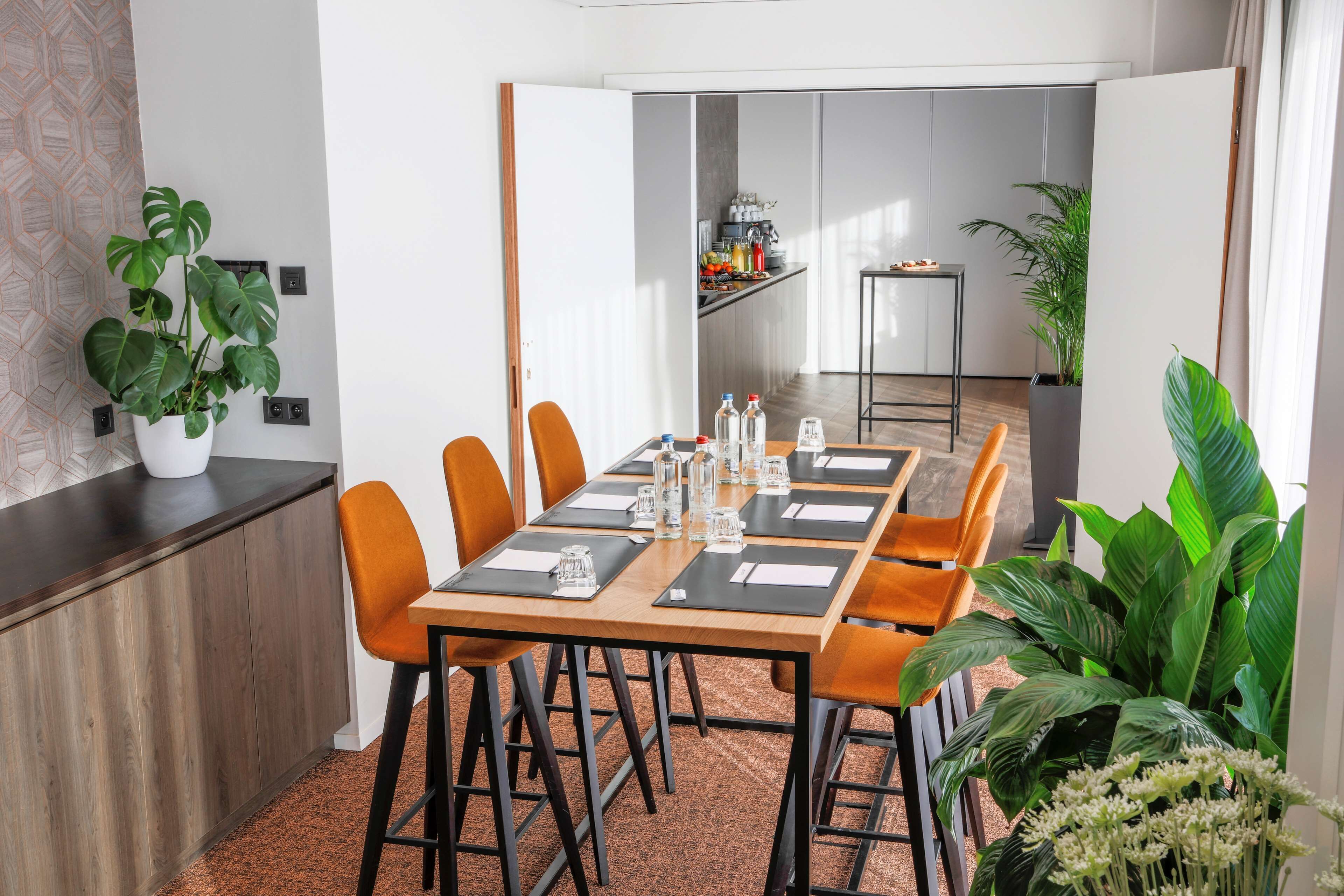 Un comedor moderno con una mesa de madera larga, sillas anaranjadas y posavasos negros. Hay plantas en macetas a los lados y una zona de cocina al fondo.