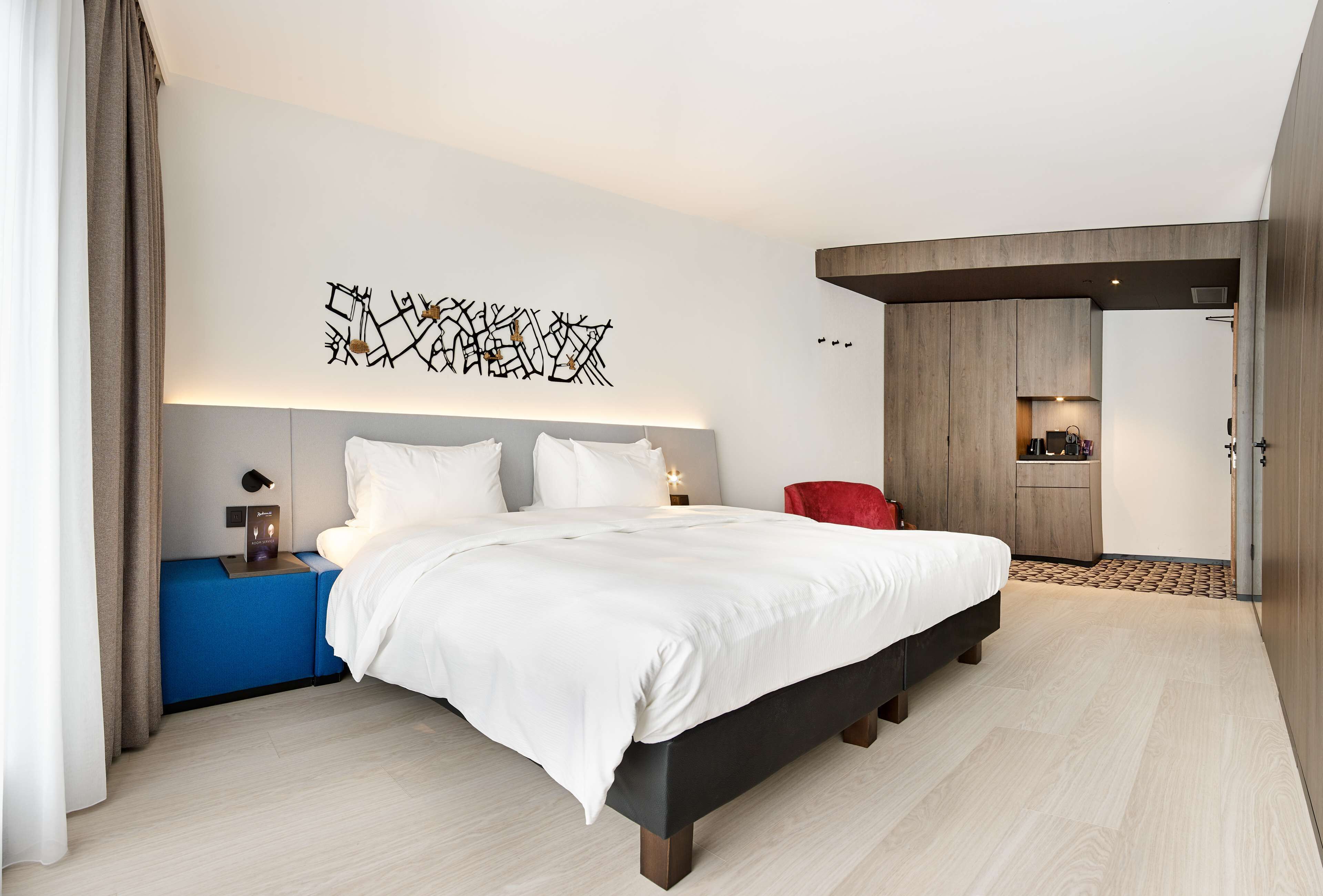 Una habitación de hotel moderna con una cama grande, una mesita de noche, una silla roja y una cocina visible a través de una puerta abierta.