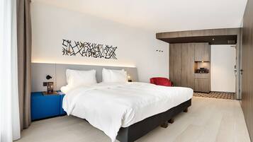 Une chambre d’hôtel moderne avec un grand lit, une table de chevet, un fauteuil rouge et un coin cuisine visible à travers une porte ouverte.