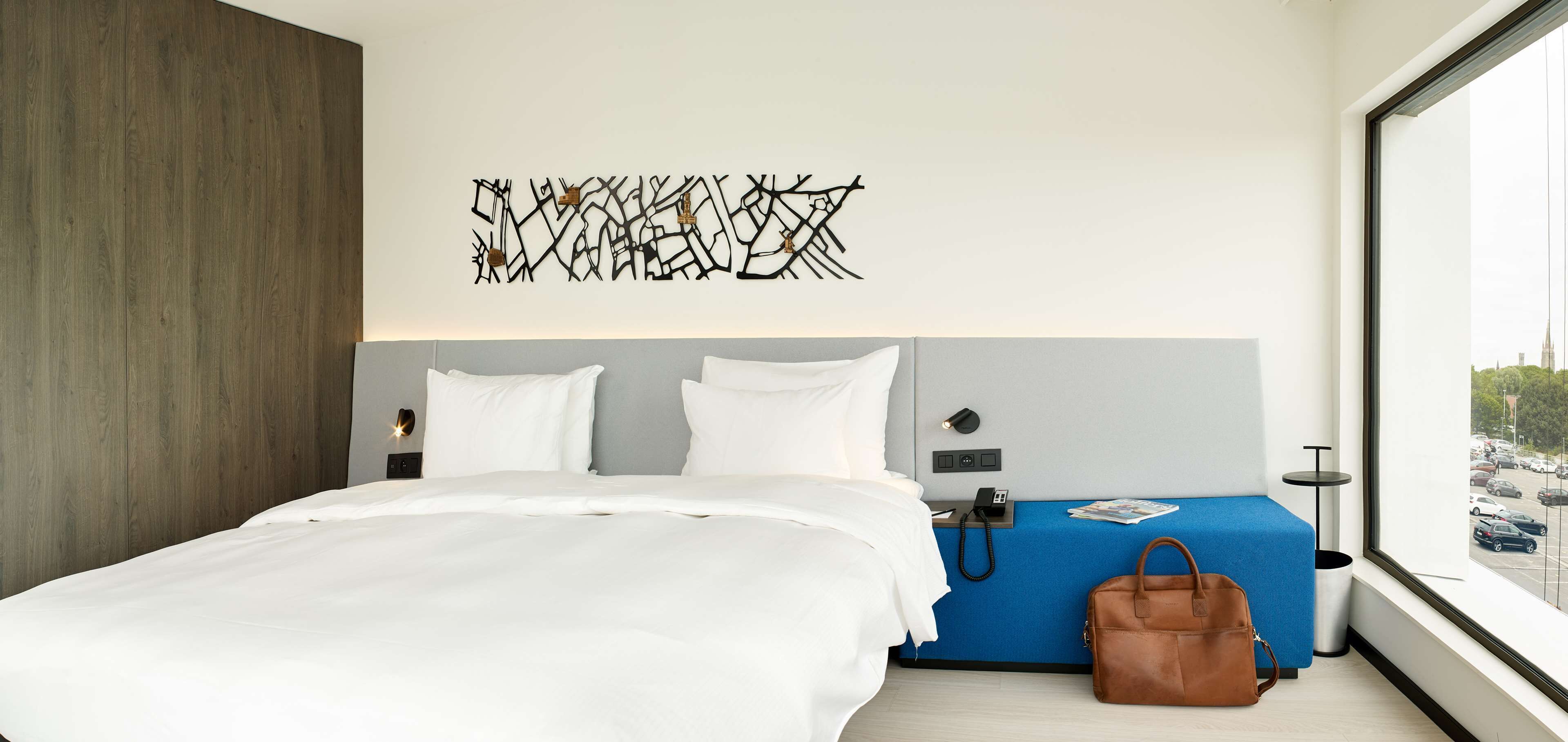 Quarto de hotel moderno com uma cama grande, um banquinho azul, uma bolsa marrom e vista para a cidade pela janela.