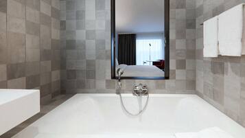 A modern bathroom with a large bathtub, a mirror, and a view of a hotel room.