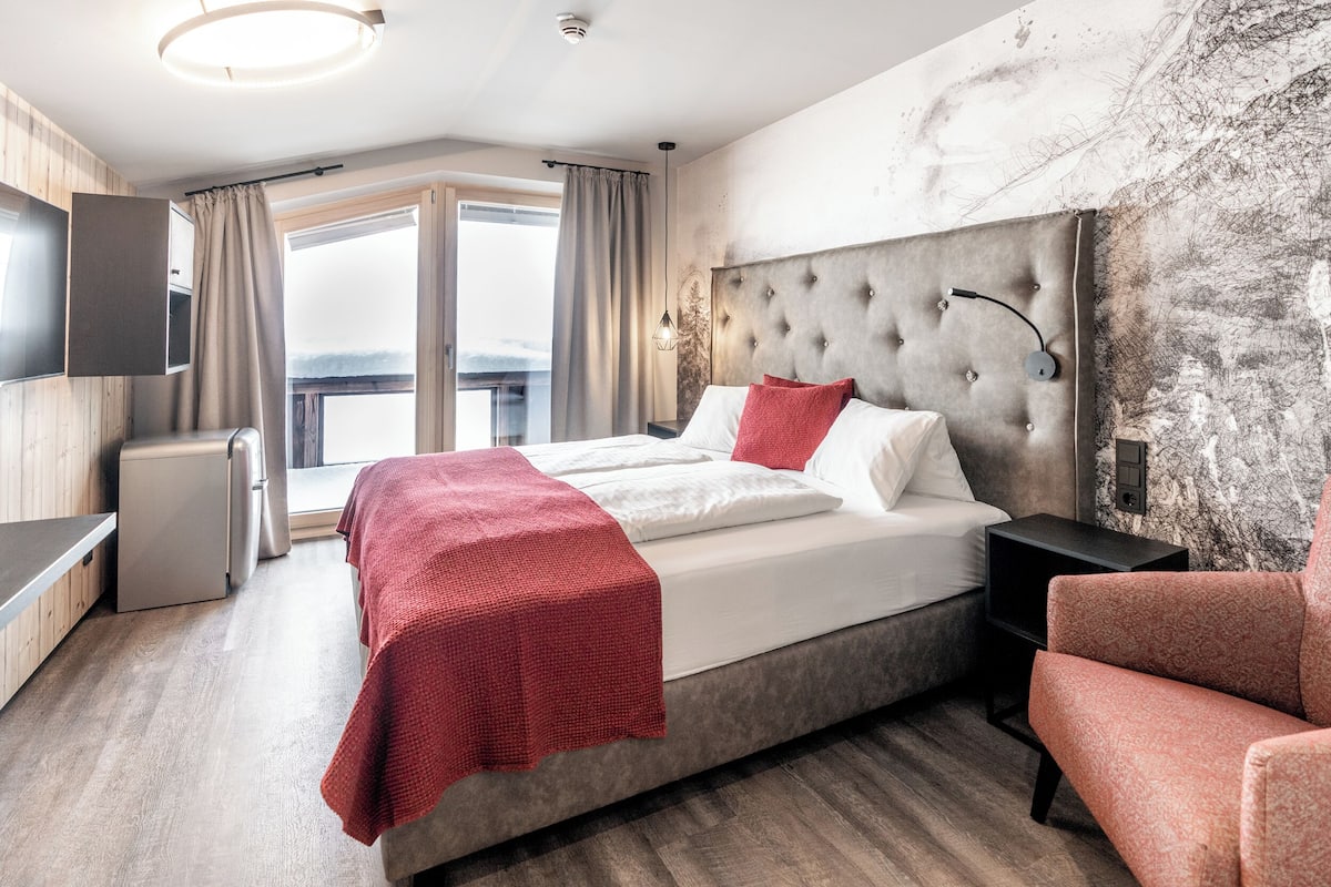A modern hotel room with a large bed, a red throw blanket, a headboard with a light fixture, a small bedside table, and a view of the outdoors through a large window.