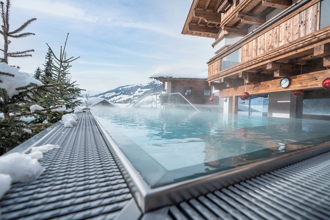 Un bâtiment en bois doté d’une piscine couverte par une verrière, entouré d’arbres et de montagnes enneigés.