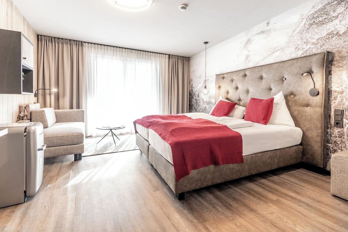 A modern hotel room with a large bed, a sofa, a desk, and a view of the outdoors.