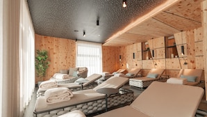 Een spa-ruimte met meerdere massagetafels, een groot raam met dunne gordijnen, en een houten wand met een stippen plafond.