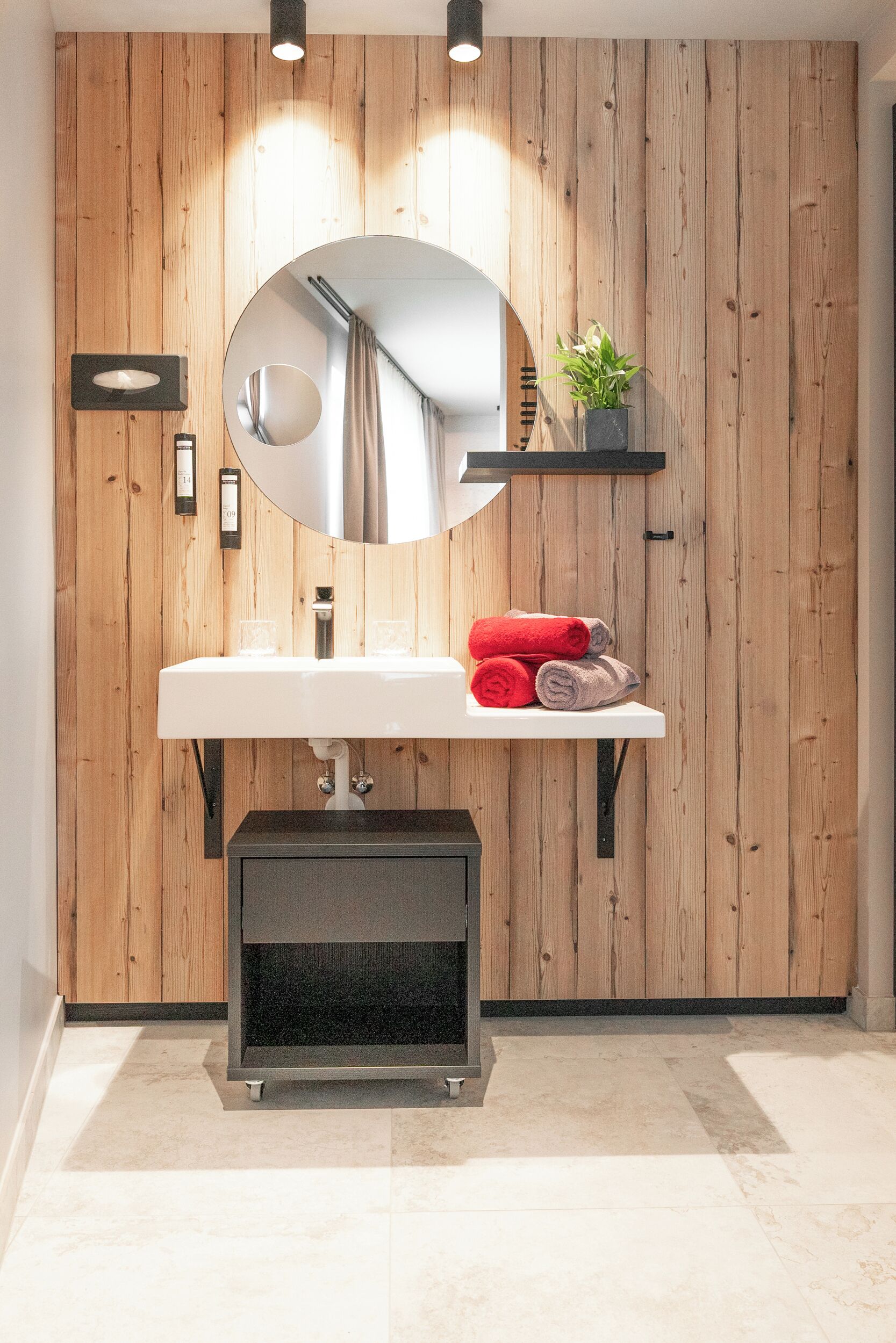 Un baño moderno con pared de madera, un lavamanos blanco, un espejo redondo y un estante pequeño con toallas y una planta.