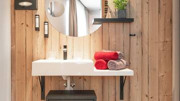 Un baño moderno con pared de madera, un lavamanos blanco, un espejo redondo y un estante pequeño con toallas y una planta.
