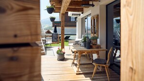 Een houten terras met buitenmeubilair, een tafel en stoelen. Er hangen plantenbakken en een bord met tekst.