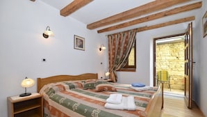A bedroom with a wooden bed, a nightstand, a lamp, a window with curtains, and a view of a stone wall through an open door. - Bella Vista Apartments (Motovun)