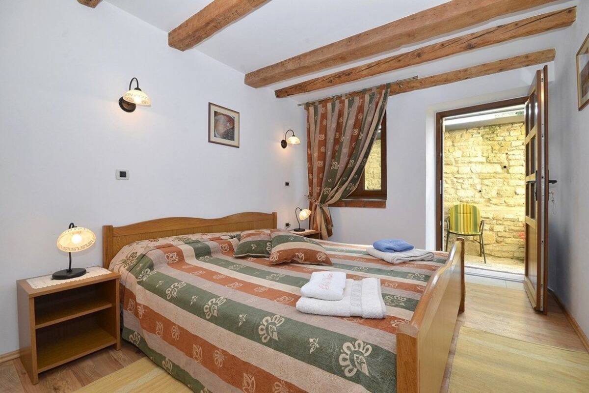 A bedroom with a wooden bed, a nightstand, a lamp, a window with curtains, and a view of a stone wall through an open door.