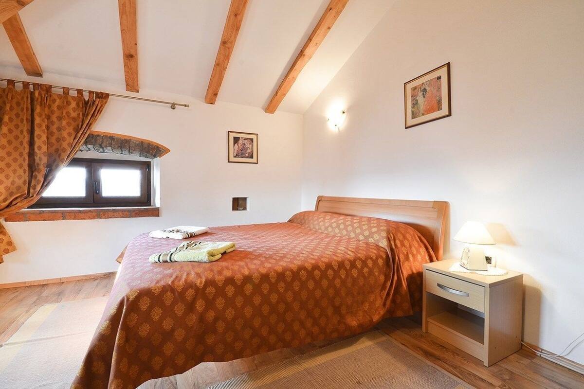 A bedroom with a wooden ceiling, a bed with a patterned bedspread, a nightstand with a lamp, and a window with curtains.