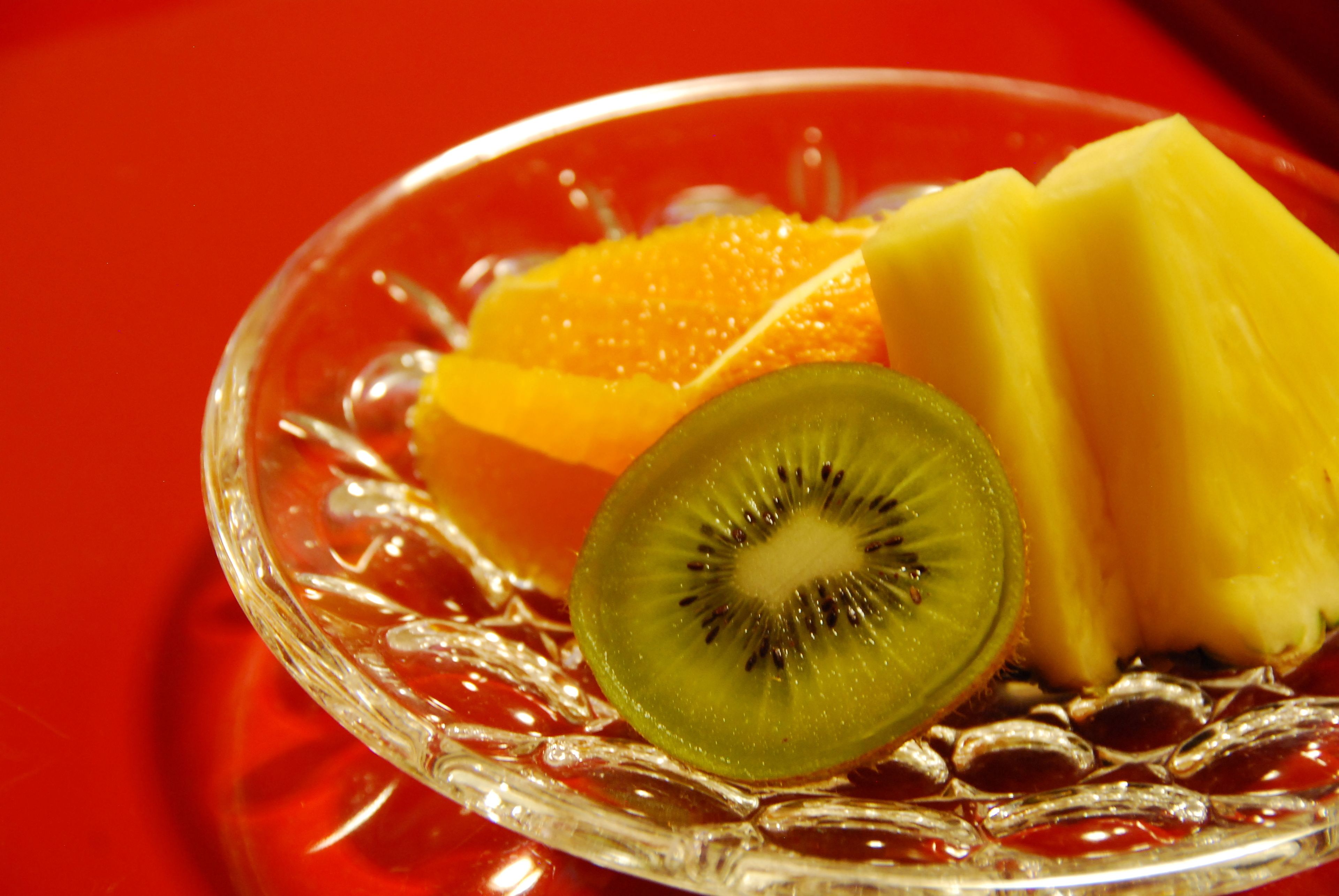 Una ciotola di vetro trasparente con una fetta di kiwi, una spicchiata di ananas e una porzione di arancia, il tutto su una superficie rossa.