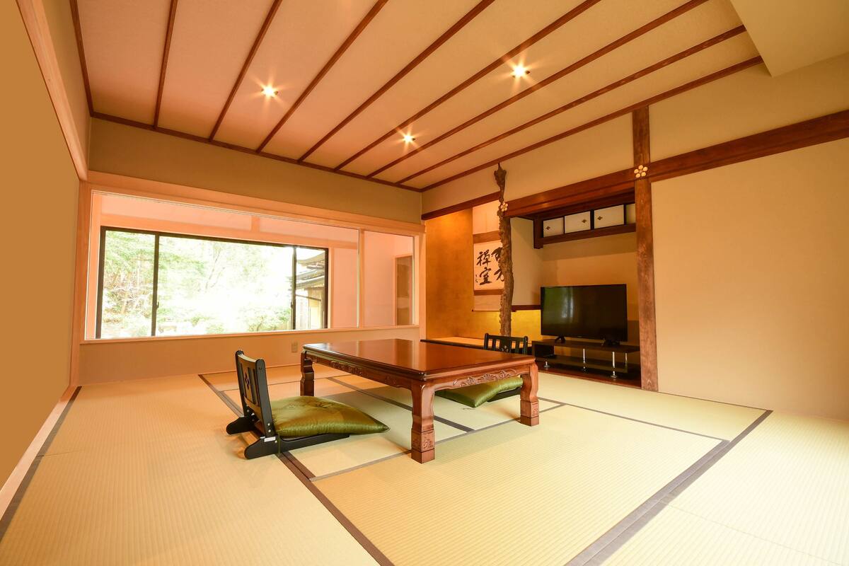 A tatami mat room with a low wooden table, green cushions, a television, and a sliding door leading to a balcony.