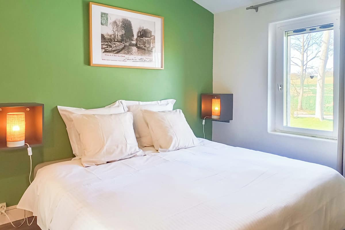 A bedroom with a green wall, a framed picture, two bedside lamps, and a window with a view of trees.