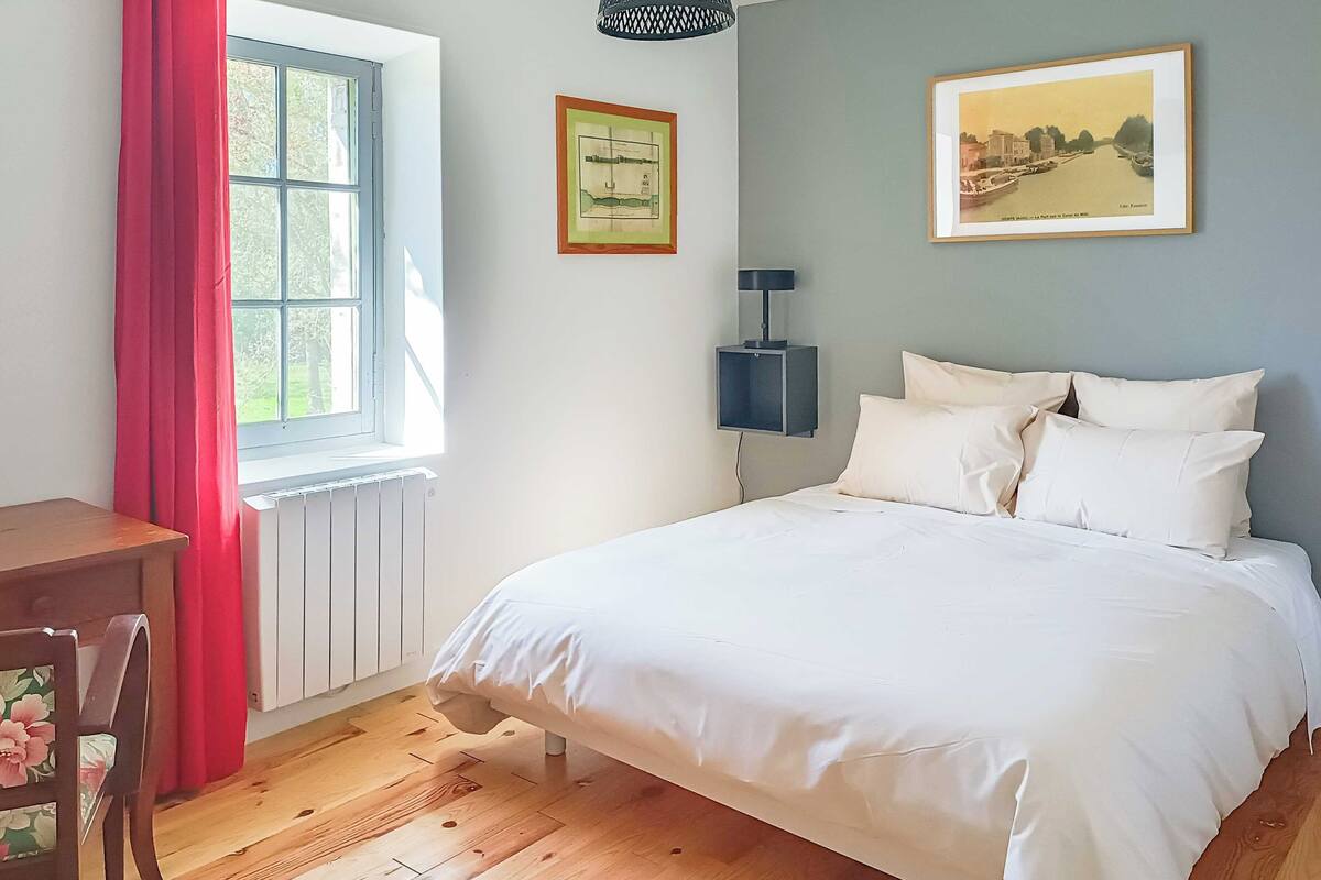 A bedroom with a large bed, a desk, a chair, a radiator, a window with curtains, and two framed pictures on the wall.