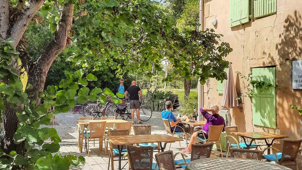 Área externa com mobiliário de vime e uma mesa, cercada por árvores e um edifício com persianas verdes.