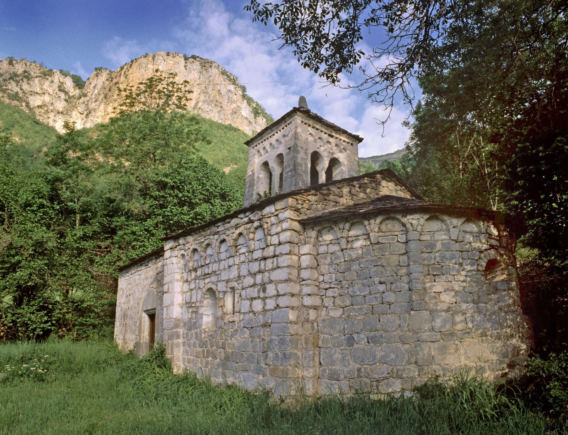 A stone church with a bell tower, surrounded by lush greenery and mountains in the background.