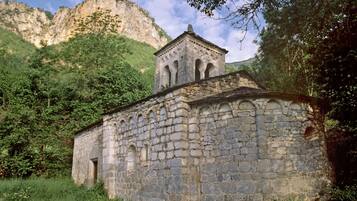 A stone church with a bell tower, surrounded by lush greenery and mountains in the background.