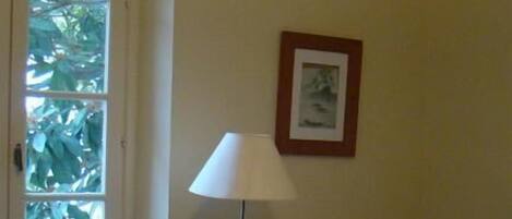 Une pièce avec un lit, un radiateur, une lampe, un tableau encadré au mur et une fenêtre donnant sur un espace verdoyant.
