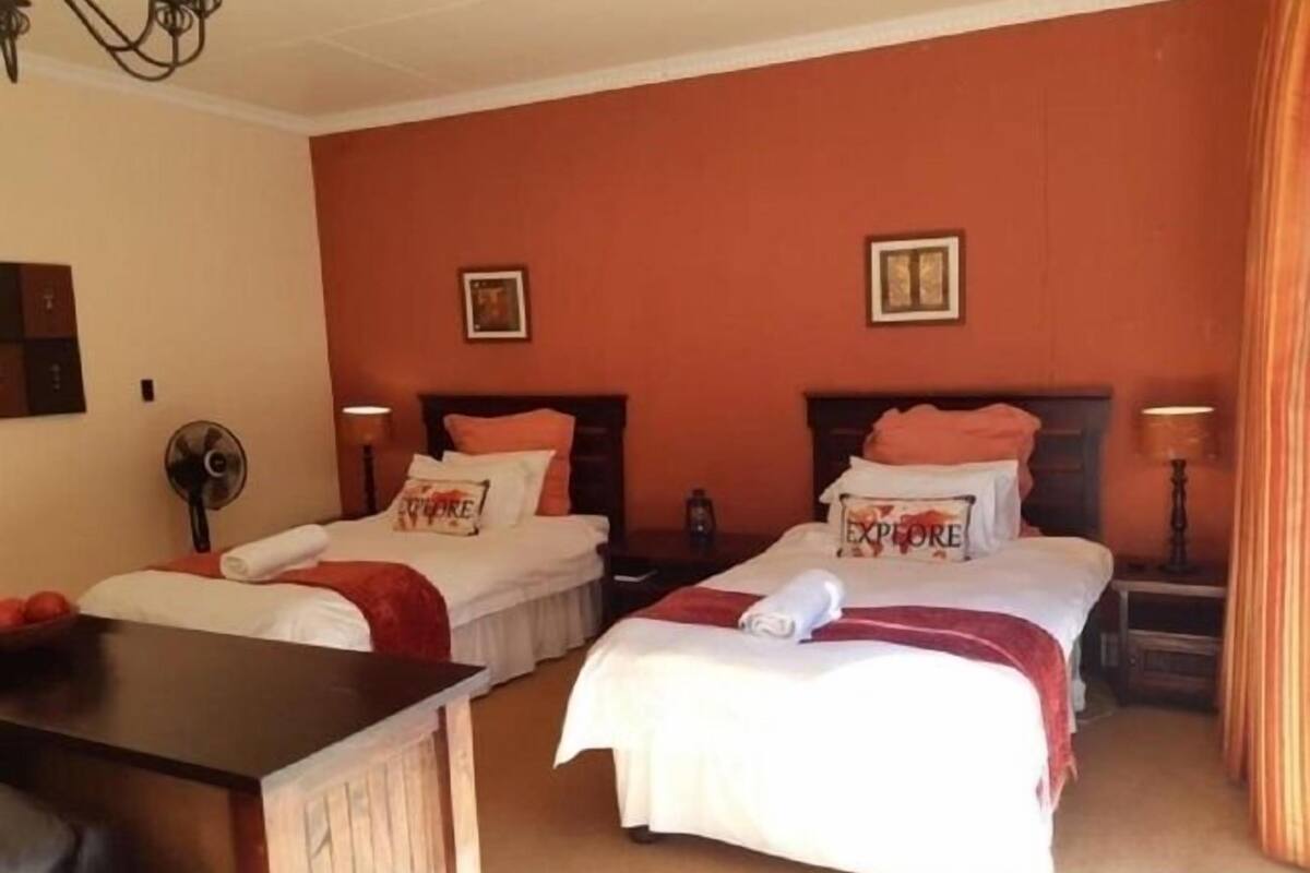 A hotel room with two beds, each with a pillow that reads "EXPLORE." There is a desk with a chair, a fan, and a lamp on each side of the room.