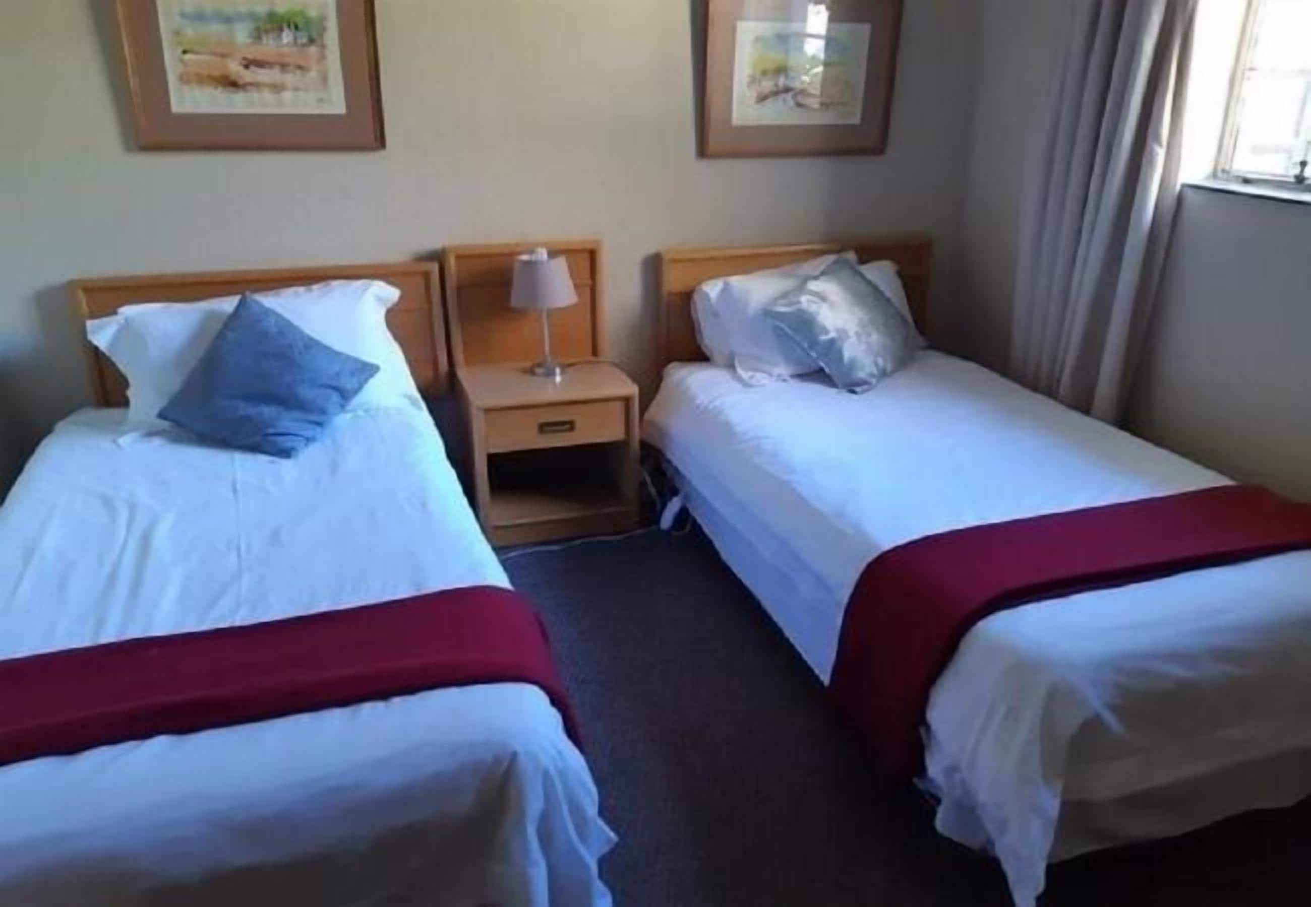 Two single beds with white bedding and red headboards, a nightstand with a lamp, and framed pictures on the wall.