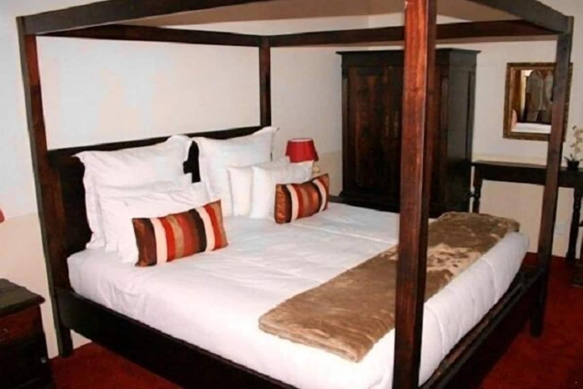 A four-poster bed with white linens and striped pillows, a wooden headboard, and a red lamp on a bedside table.