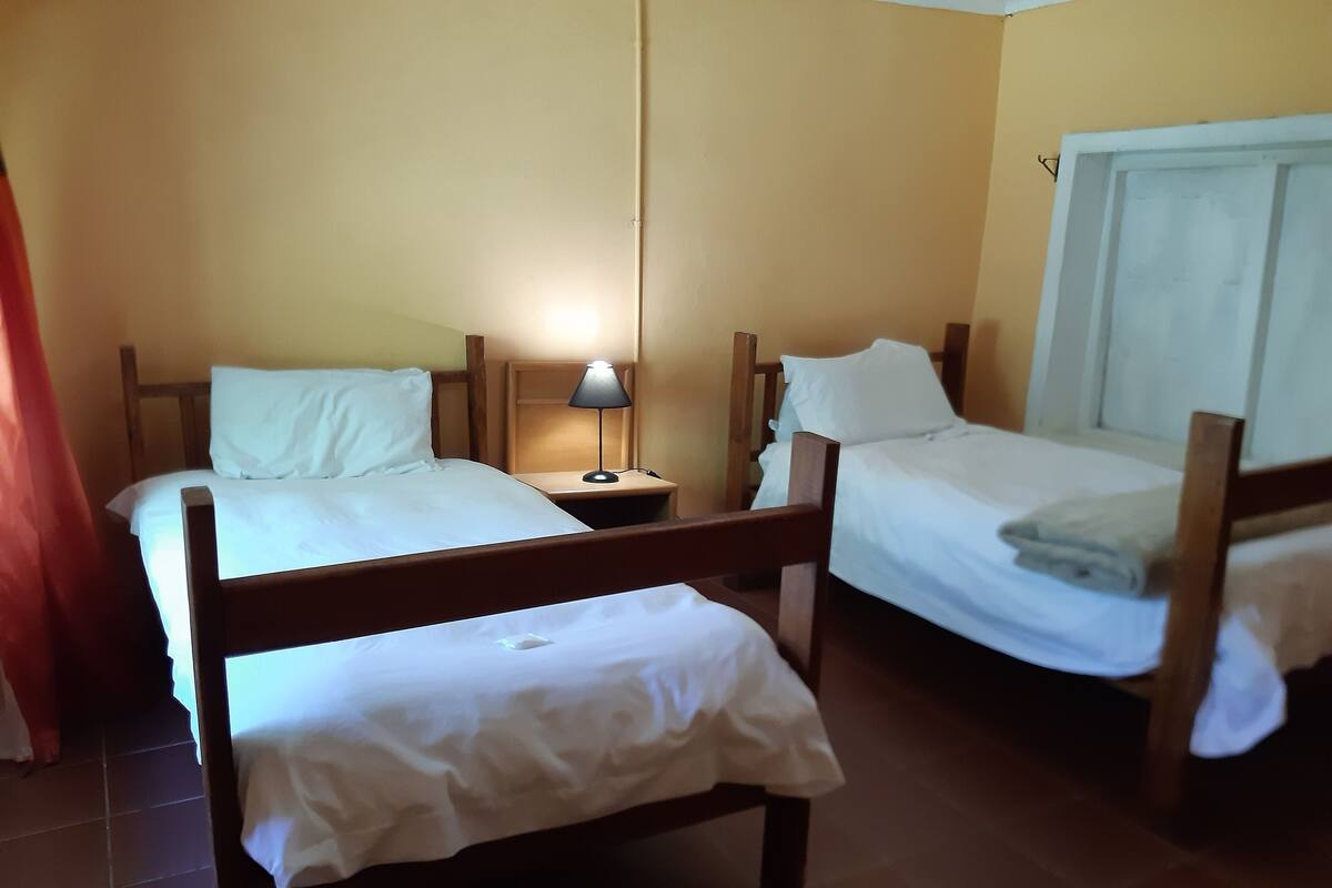A room with two wooden beds, each with white bedding and pillows, a nightstand with a lamp, and a window with curtains.