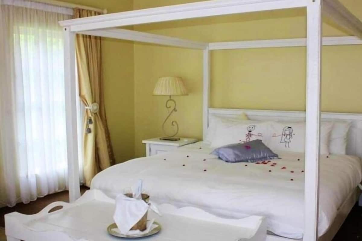 A four-poster bed with white bedding, a bedside table with a lamp, and a window with curtains.