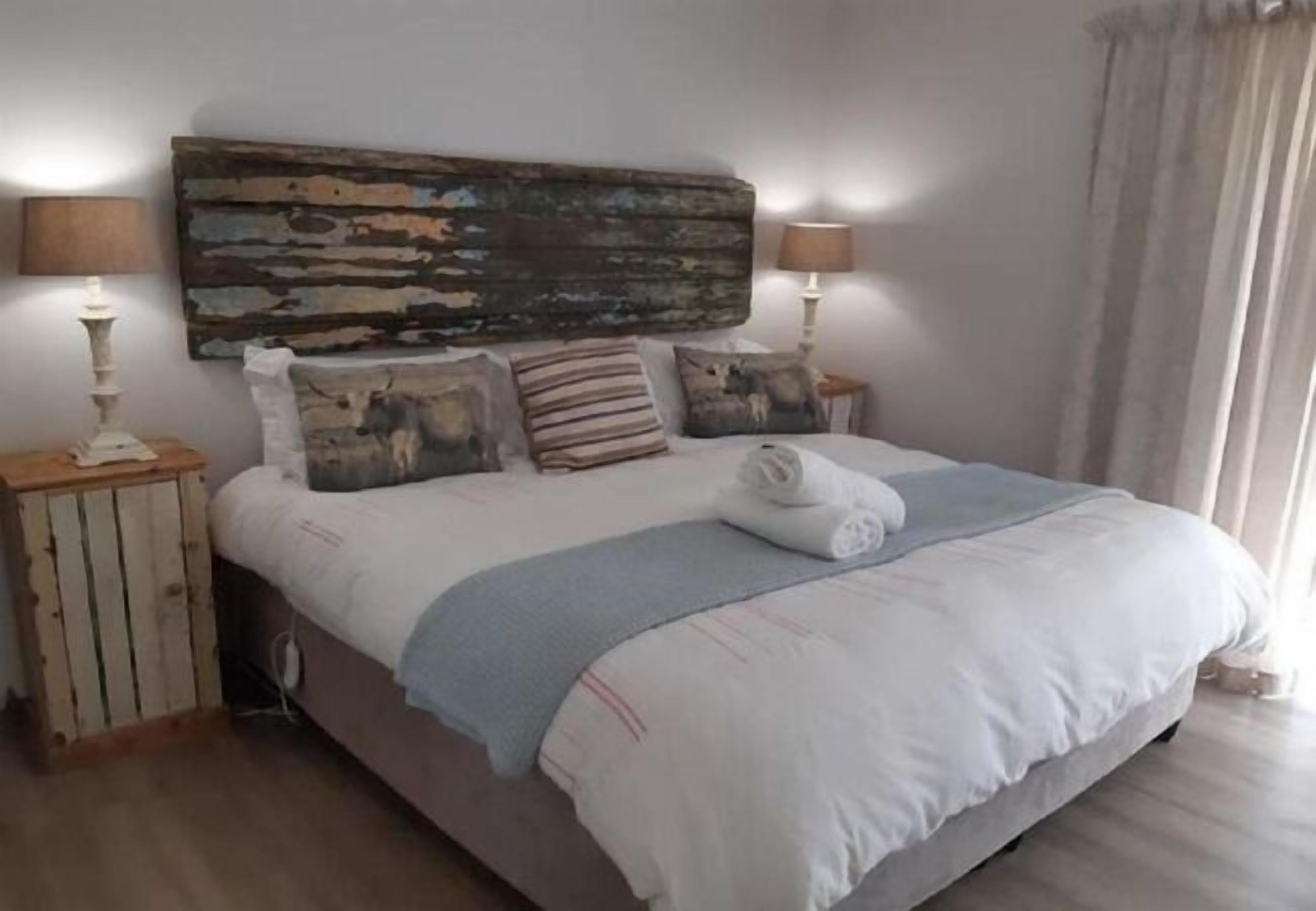 A neatly made bed with a white comforter and a blue throw blanket, flanked by two bedside tables with lamps, a rustic headboard, and a large abstract painting above the bed.