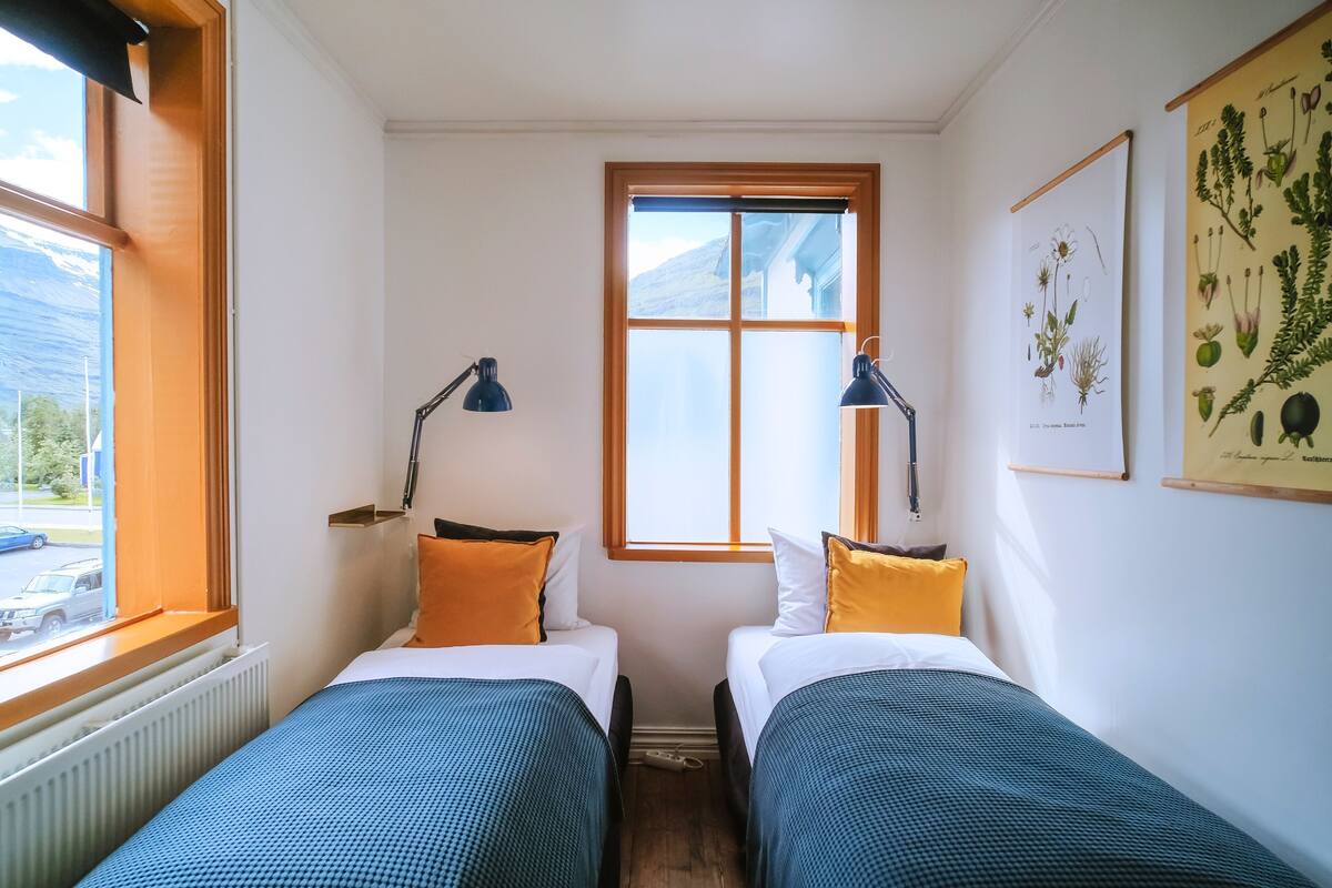 A small, well-lit room with two single beds, a window with a view of the outside, and a wall-mounted plant illustration.