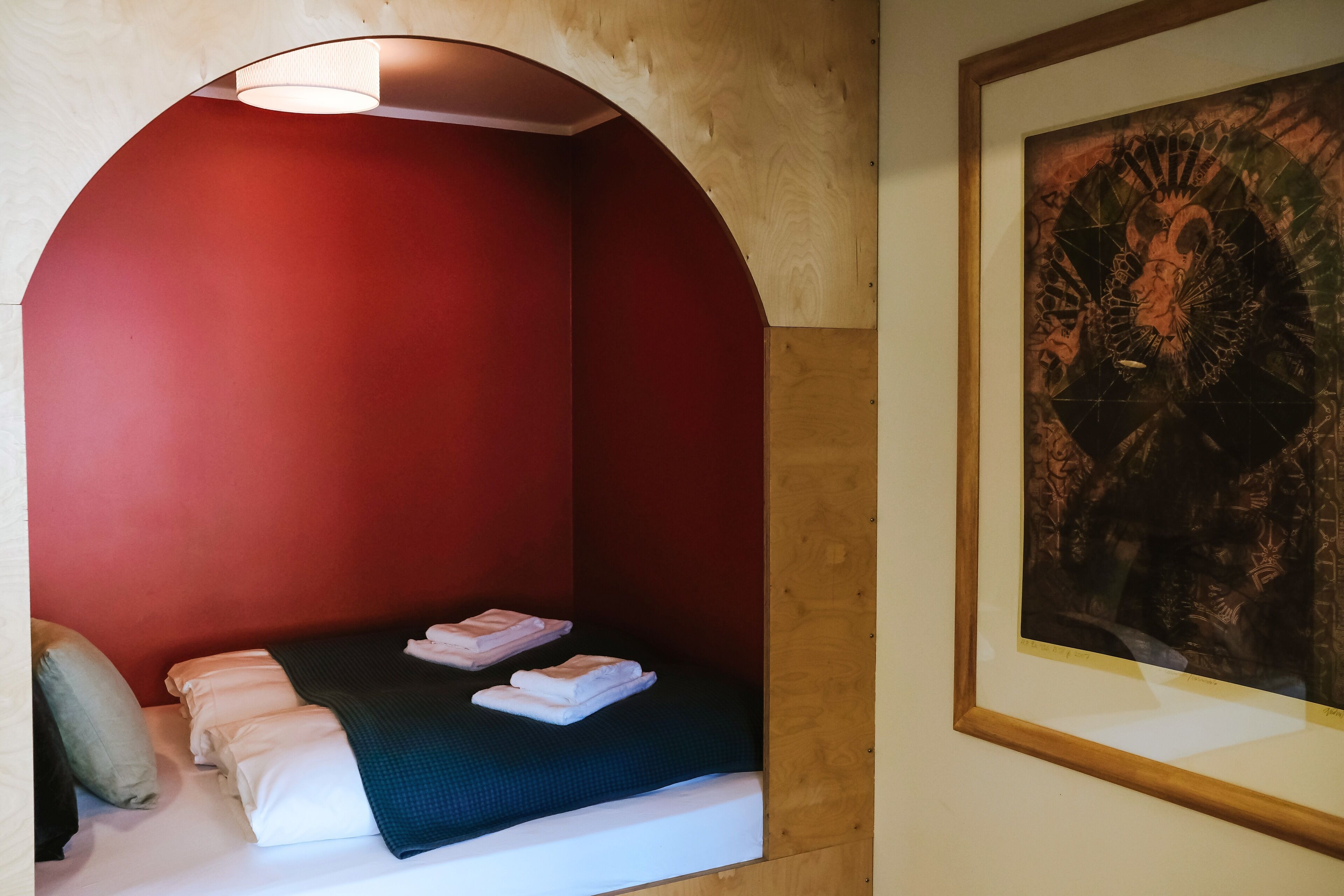 Une petite pièce agréable et chaleureuse, avec un lit simple, un mur rouge et une œuvre d’art encadrée accrochée au mur.