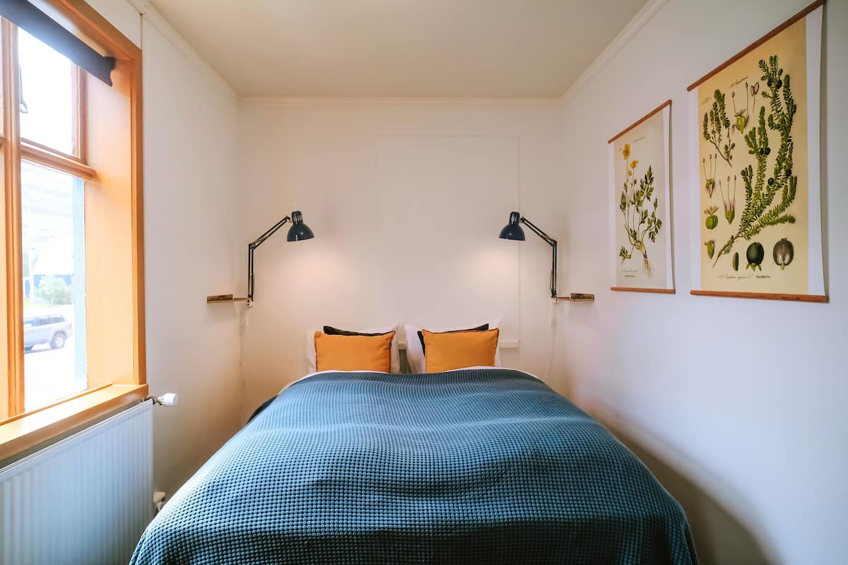 A bedroom with a bed, two bedside lamps, a window, and two botanical posters on the wall.