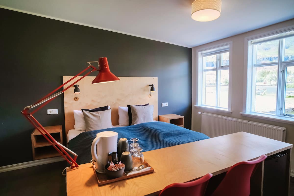 A bedroom with a bed, bedside tables, a desk with chairs, a red desk lamp, and a window with a view.