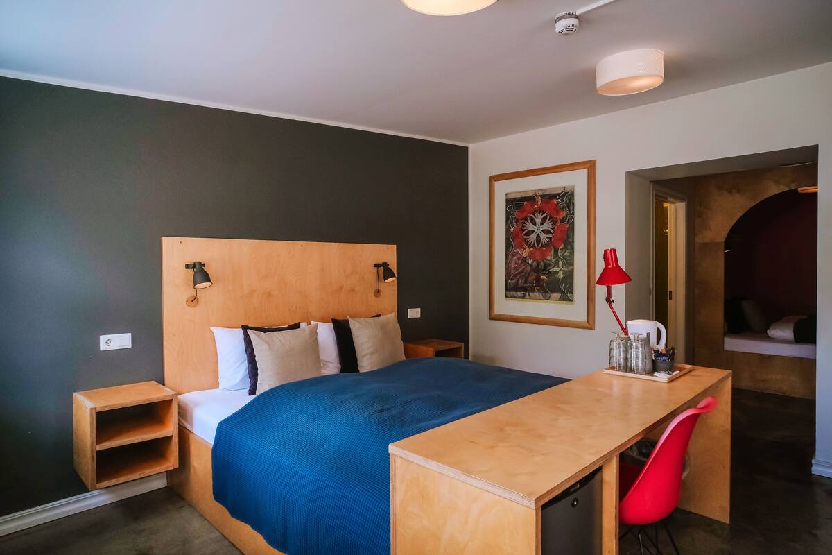 A modern bedroom with a wooden headboard, a blue bedspread, a wooden desk, and a red chair.