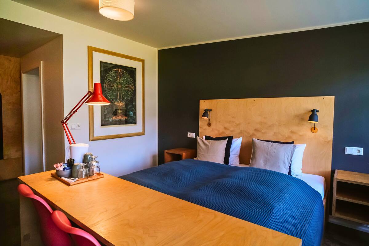 A bedroom with a wooden headboard, a bed with a blue bedspread, a red desk lamp, a framed picture on the wall, and a small wooden nightstand.