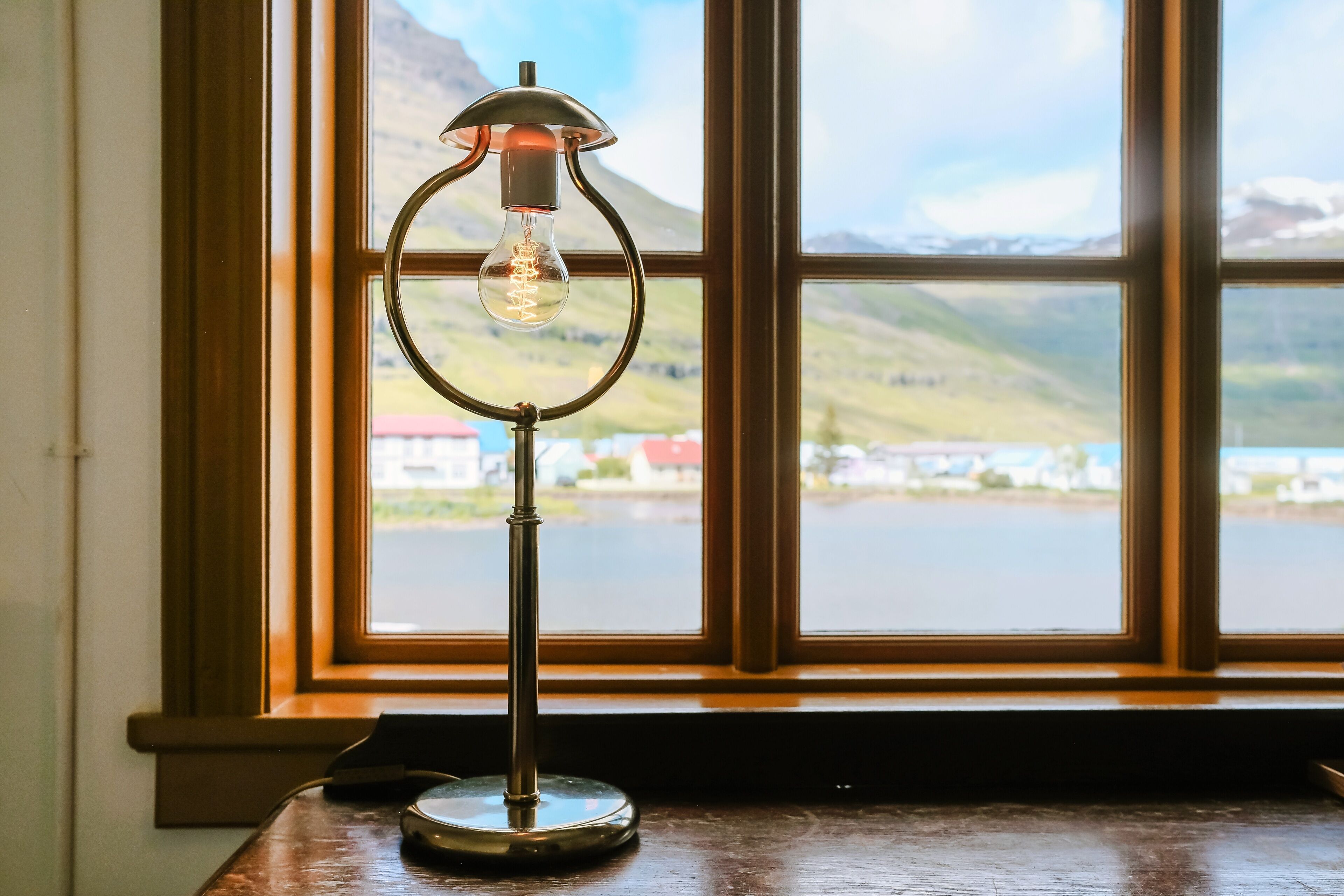 Une lampe de table avec une ampoule visible, posée sur une table en bois près d’une fenêtre donnant sur un lac et des collines.