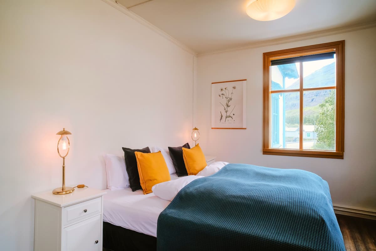 A neatly made bed with a blue blanket, white pillows, and a framed botanical print on the wall.