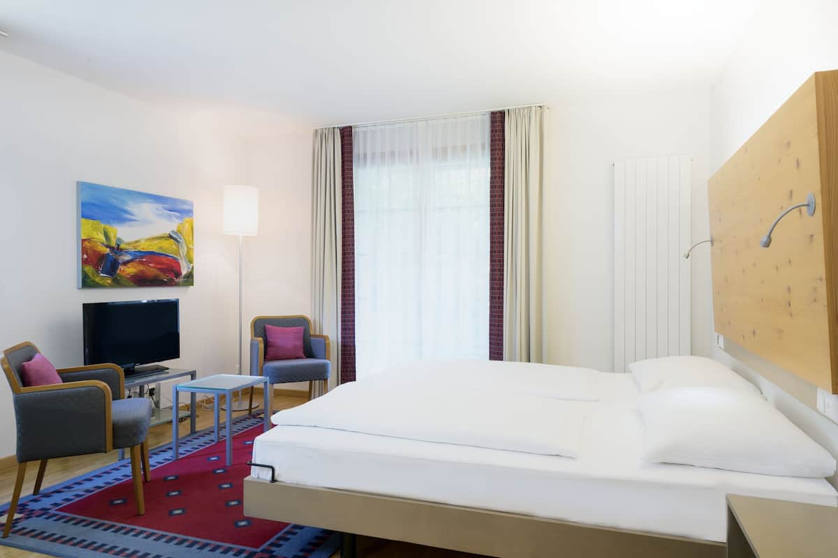 A hotel room with a bed, a TV, a desk, two chairs, a colorful rug, and a painting on the wall.