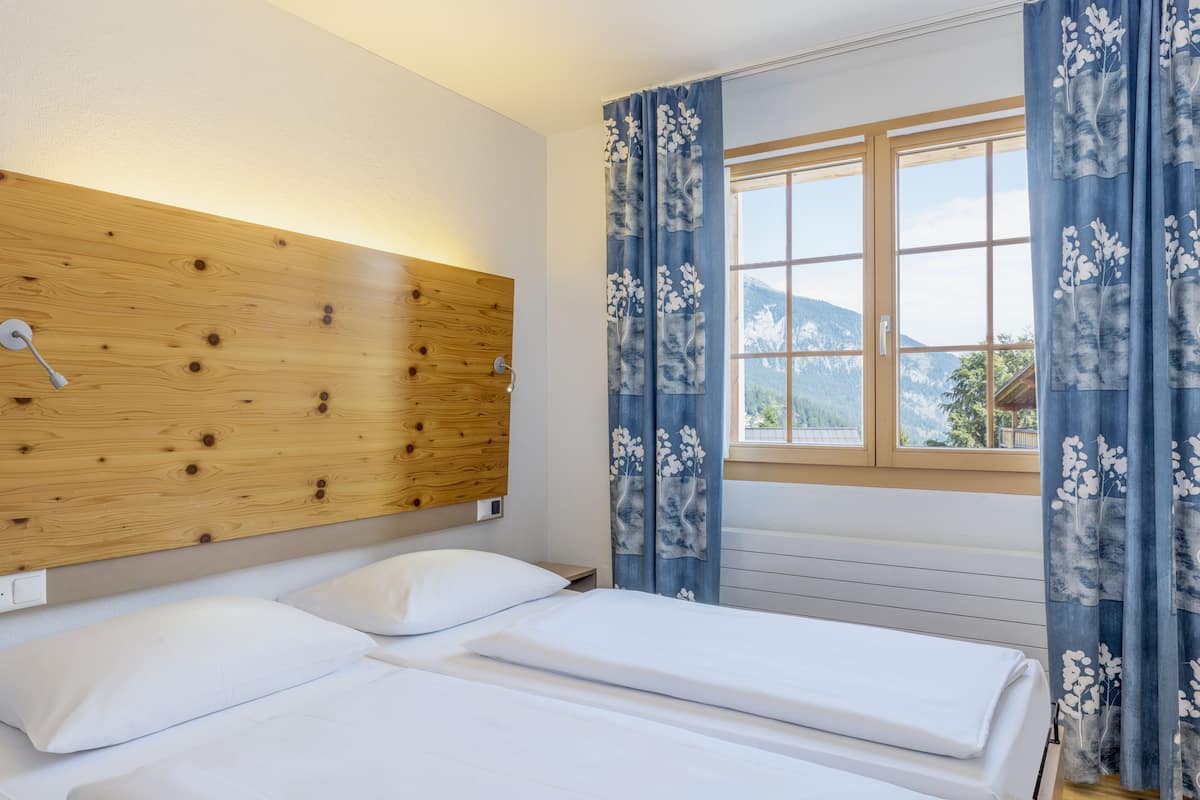 A bedroom with two beds, a wooden headboard, a window with curtains, and a view of mountains.