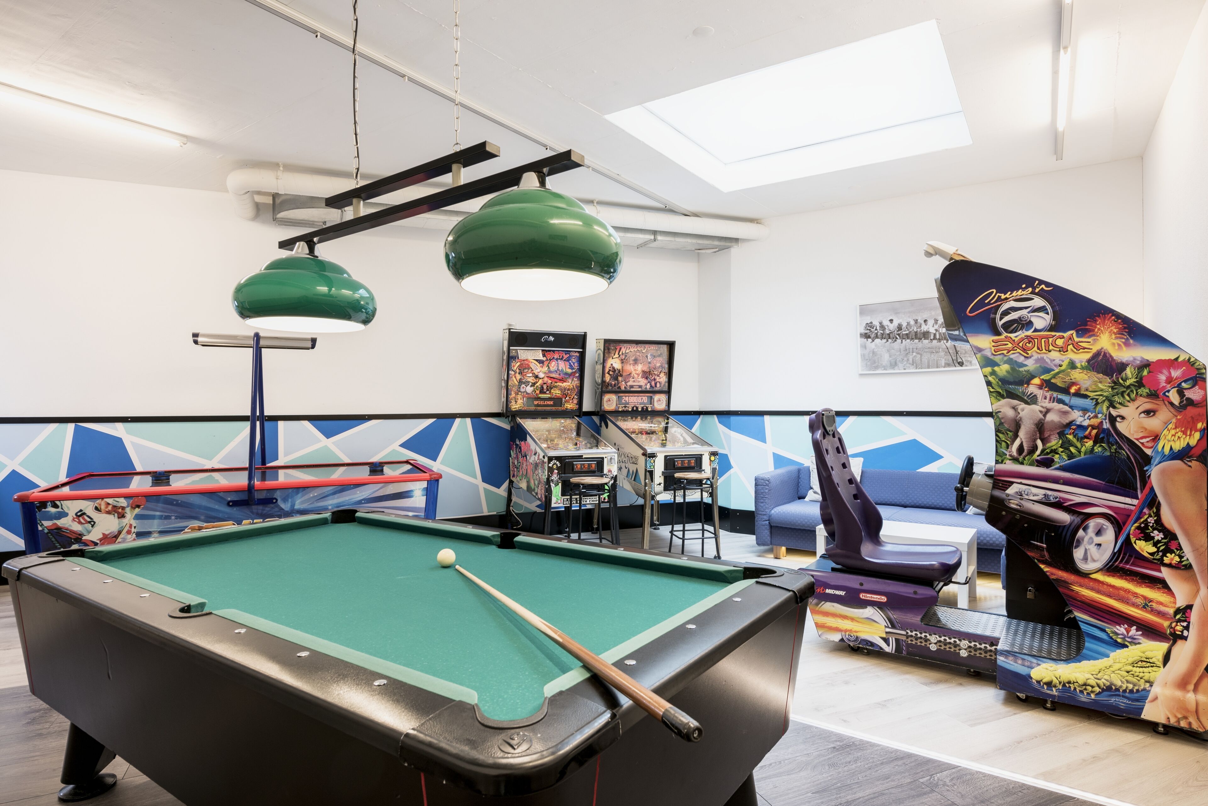 A pool table with a pool cue, a green overhead lamp, arcade games, and a blue chair.
