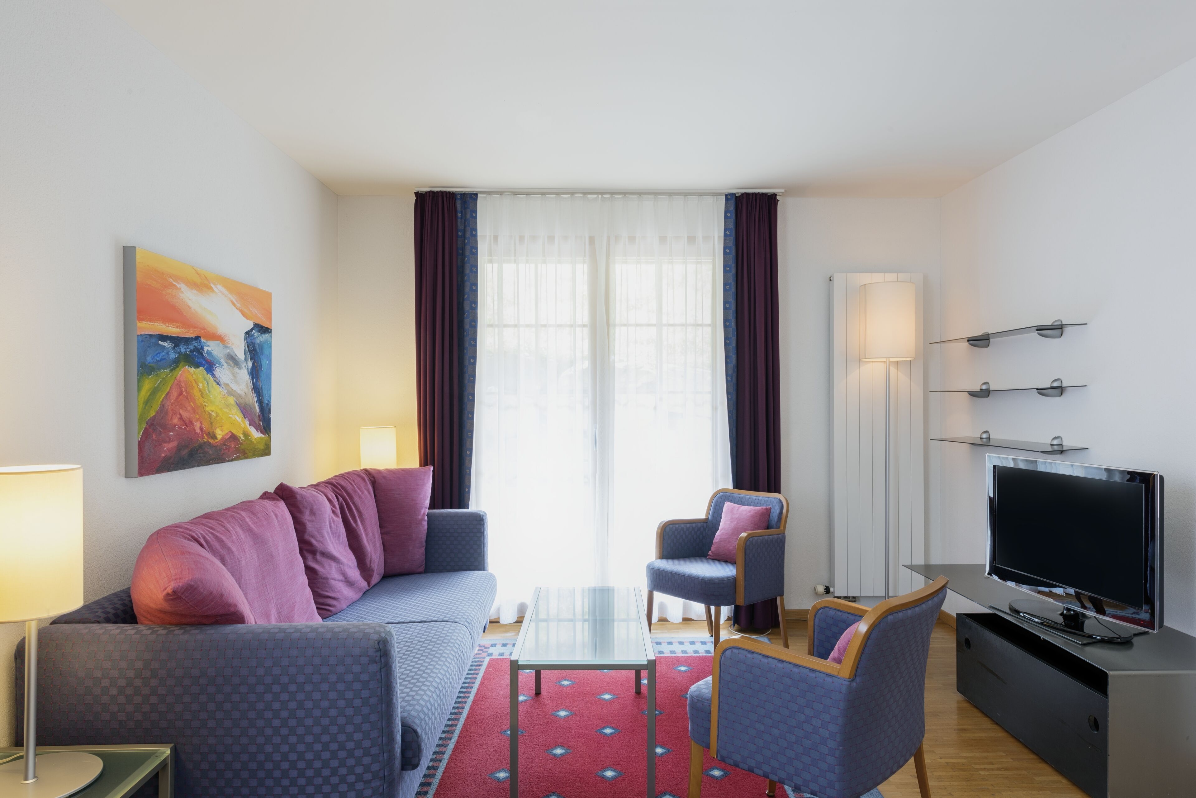 A modern living room with a sofa, armchairs, a glass coffee table, a flat-screen TV, and a painting on the wall.