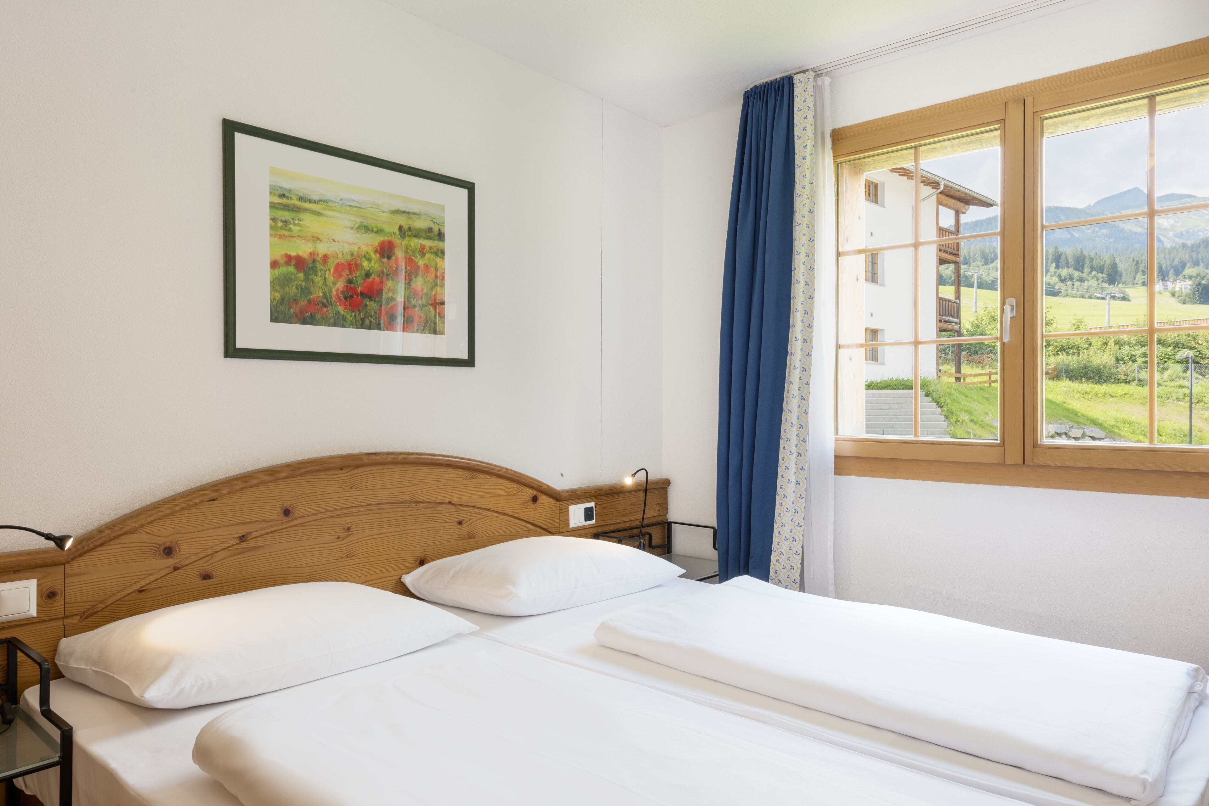 Ein Hotelzimmer mit einem Bett mit Holz-Kopfteil, einem gerahmten Landschaftsbild und einem Fenster mit Blick auf ein Gebäude und Grünflächen.