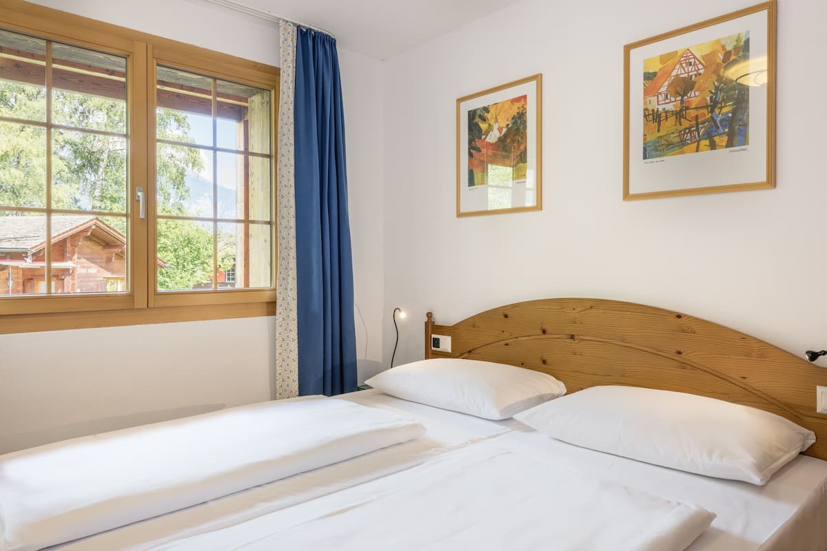 A hotel room with two single beds, a wooden headboard, a window with blue curtains, and two framed artworks on the wall.
