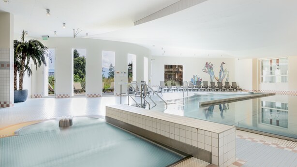An indoor swimming pool with clear glass walls, a tiled floor, and a seating area with chairs and a palm tree. - Hapimag Resort Flims (Flims)