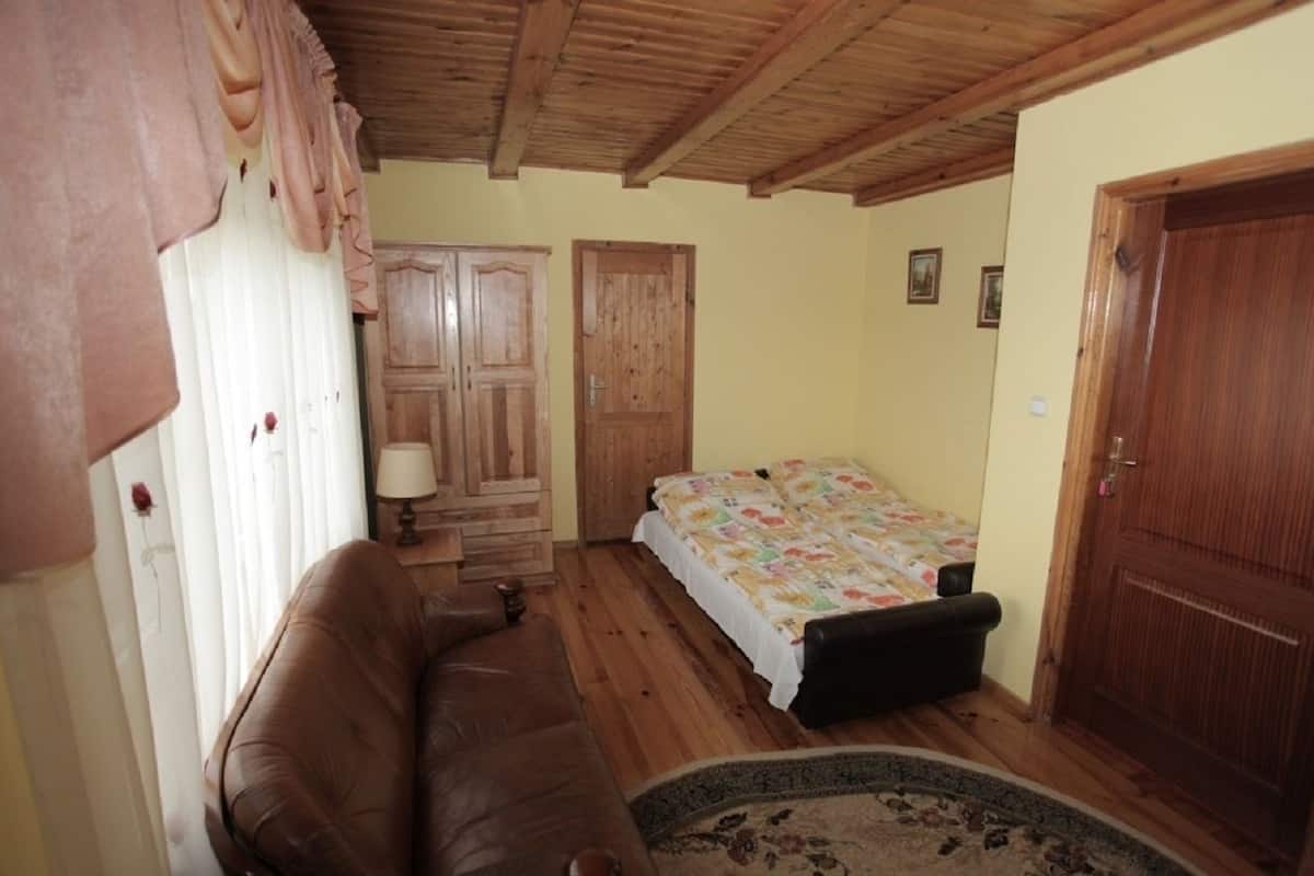 A small, cozy room with a wooden ceiling, a brown leather sofa, a single bed with a floral bedspread, a wooden wardrobe, and a door.