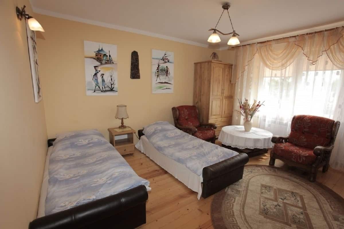 A room with two single beds, a wooden wardrobe, a red armchair, a small round table with a vase, and a large window with curtains.